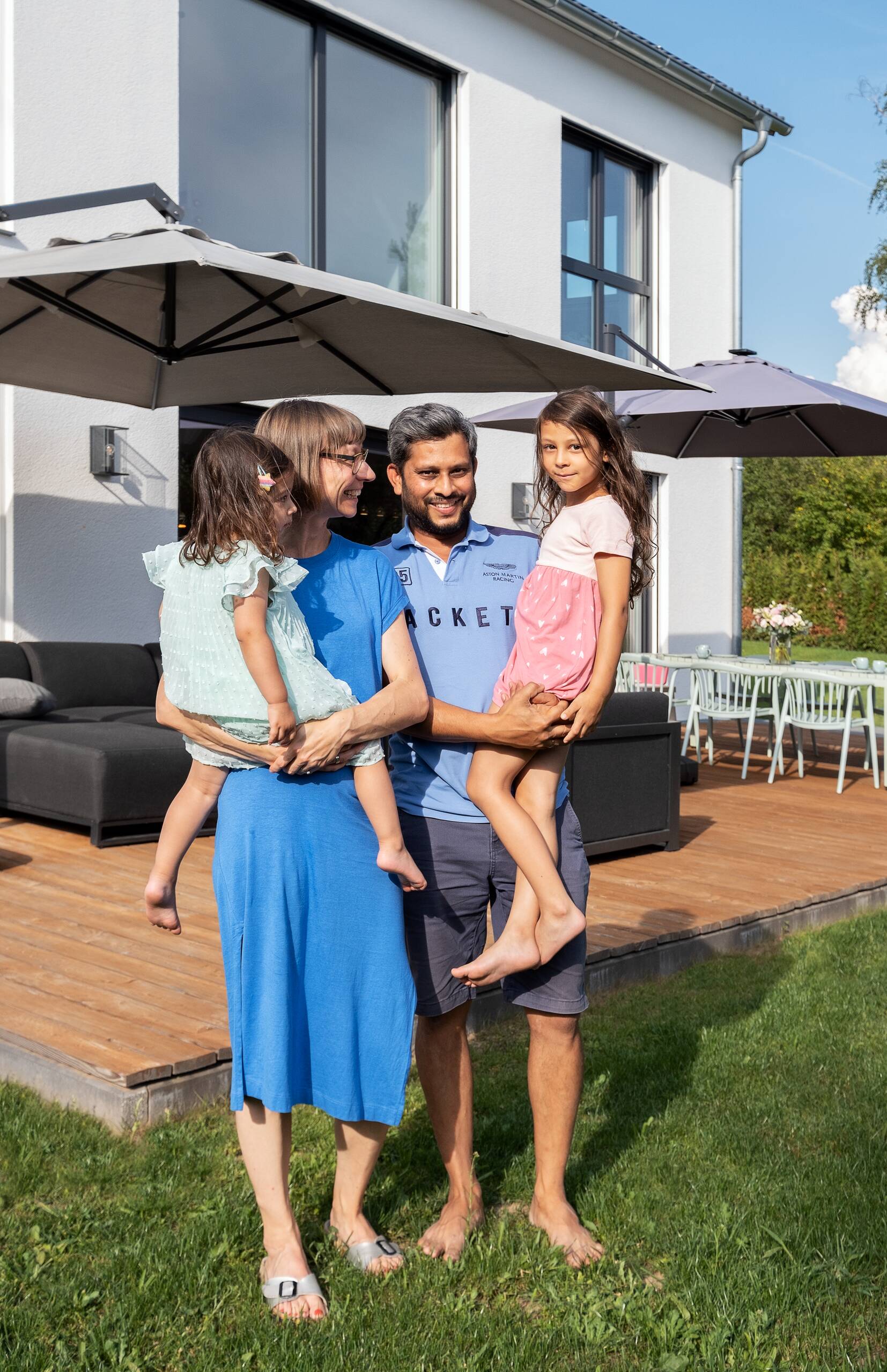 Eine Familie steht im Garten eines Hauses. Ein Mann hält ein Kind auf dem Arm, während eine Frau ein weiteres Kind trägt. Alle lächeln und wirken entspannt. Der Garten ist mit einer Holzterrasse und einem Essbereich ausgestattet.