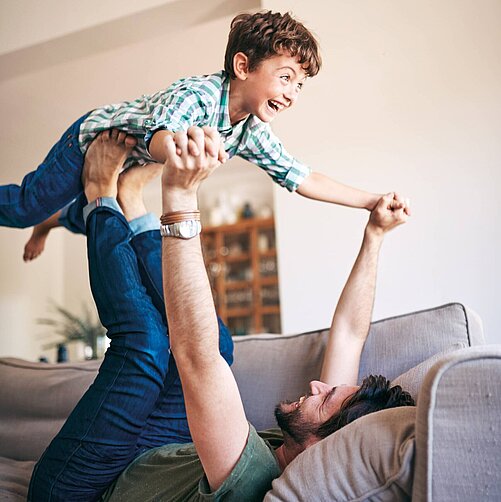 Ein Vater liegt auf einem Sofa und hebt seinen fröhlichen Sohn in die Luft. Der Junge lacht und freut sich, während er von den Händen des Vaters gehalten wird. Es strahlt ein Gefühl von Freude und Verspieltheit aus.