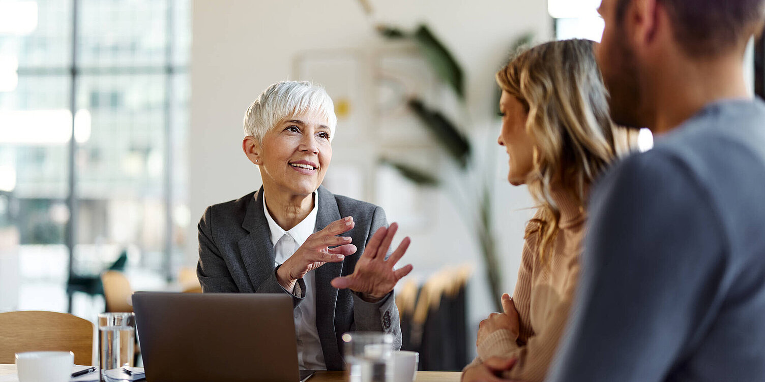 Eine Beraterin mit kurzem grauen Haar und einem freundlichen Lächeln spricht mit einem jungen Paar in einem modernen Büro. Vor ihr steht ein aufgeklappter Laptop, die Atmosphäre wirkt professionell und zugleich entspannt.
