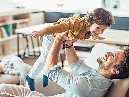 Ein Vater hält seinen fröhlichen Sohn in der Luft, während sie miteinander lachen. Der Junge trägt ein gelbes Oberteil und hat seine Arme ausgebreitet, als ob er fliegt. Die Szene strahlt Freude und Liebe in einem gemütlichen Wohnraum aus.