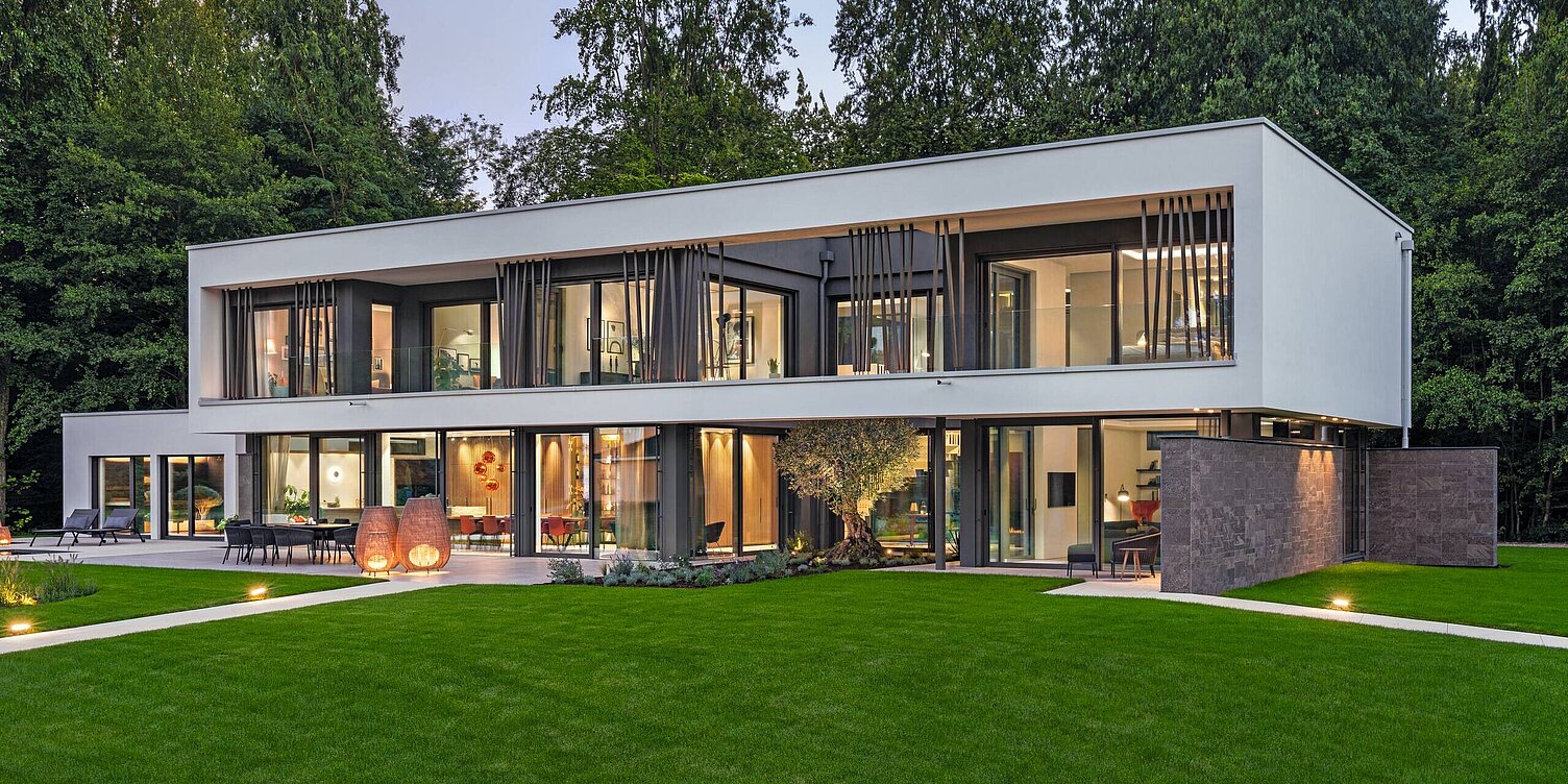 Ein modernes, zweistöckiges Haus mit großen Fensterfronten und einem grünen Garten. Das Gebäude hat eine minimalistische Architektur, umgeben von Bäumen. Die Fassade ist weiß und es gibt einen gepflasterten Bereich mit dekorativen Elementen im Vordergrund.