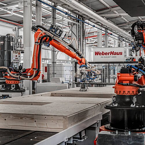Fertighausproduktion bei WeberHaus: In einer modernen Produktionshalle sind mehrere orangefarbene Industrieroboter zu sehen, die an einer Fertigbauwand arbeiten. Im Hintergrund befindet sich ein Schild mit der Aufschrift „WeberHaus – Die Zukunft leben“. Die Umgebung ist hell und industriell gestaltet.