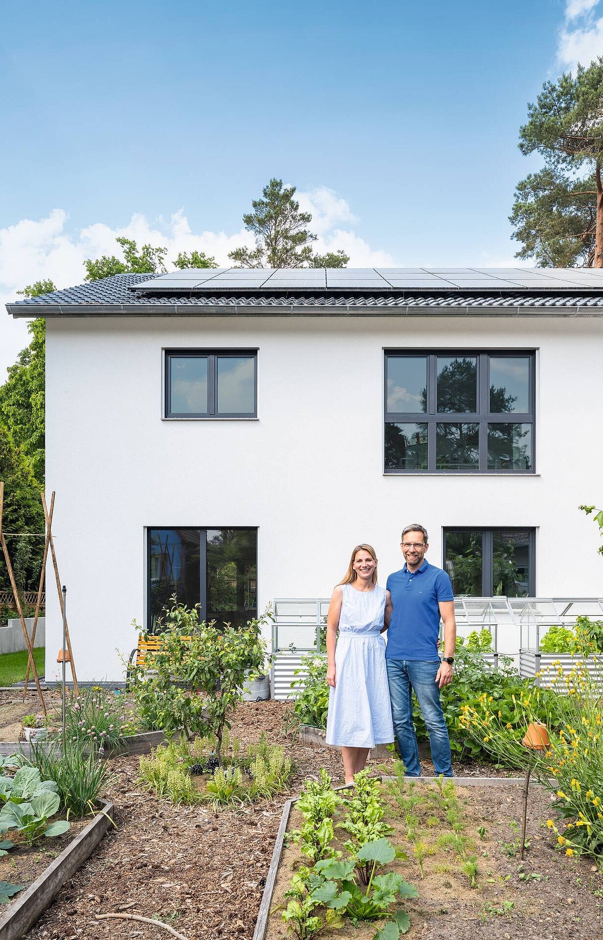 Ein Paar steht in einem Garten vor ihrem modernen, weißen Haus. Der Garten ist mit Gemüsebeeten und bunten Blumen angelegt. Im Hintergrund sind Bäume und ein blauer Himmel sichtbar. Die Personen lächeln und strahlen Freude aus.